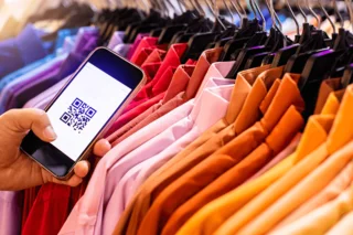 vor einer mit Textilien gefüllten Kleiderständer wird ein Smartphone mit einem QR-Code gehalten